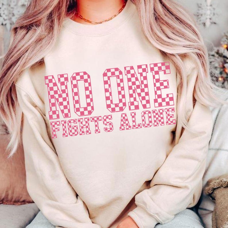 May include: A beige sweatshirt with the text "NO ONE FIGHTS ALONE" in a pink checkered pattern. The sweatshirt has a crew neck and long sleeves. The text is centered on the front of the shirt.