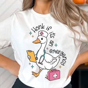 Puede incluir: Camiseta blanca con un pato de dibujos animados que lleva un gorro de enfermera y sostiene un portapapeles y un estetoscopio. El texto "Honk if it's an emergency" est&aacute; impreso en la camiseta.