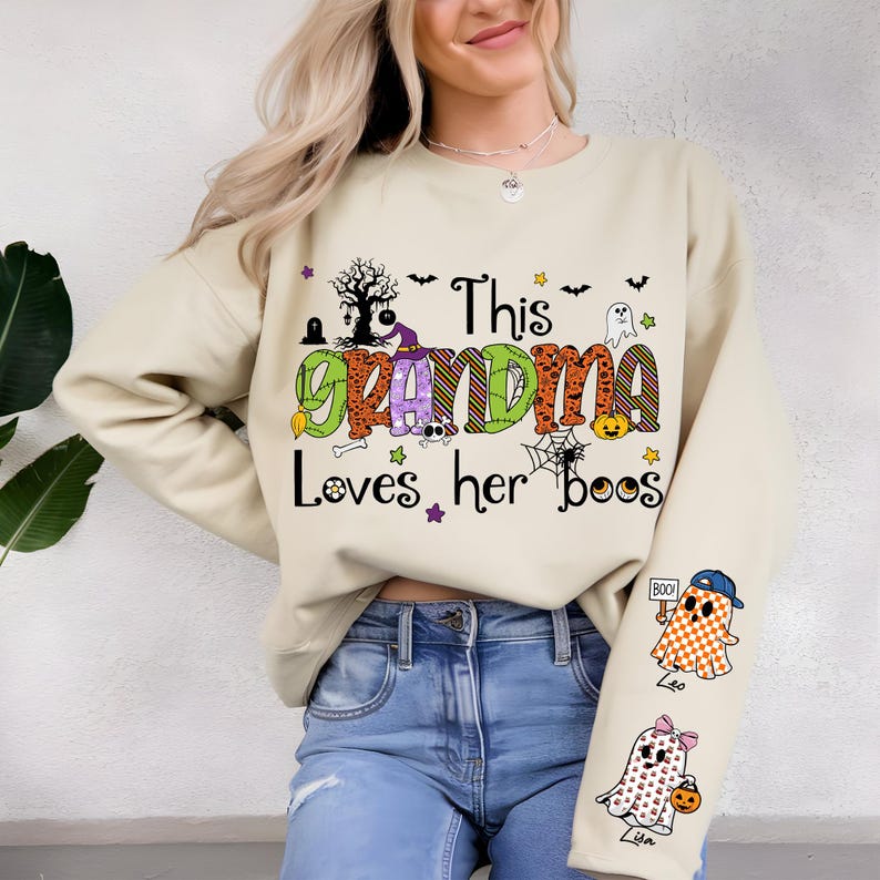May include: Beige sweatshirt with the text "This Grandma Loves her boos" in green, orange, and purple. The design includes a tree, bats, ghosts, and pumpkins. The sleeves feature ghost illustrations with names.