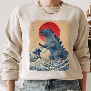 May include: Beige sweatshirt with a Japanese-inspired Godzilla design. The artwork features a red sun, blue waves, and the word "Kaiju" in Japanese. The graphic has a distressed, vintage appearance, reminiscent of traditional woodblock prints.