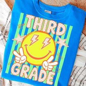 May include: A bright blue sweatshirt with a graphic design for third grade. The design features a yellow smiley face with lightning bolt eyes, peace signs, stars, and the words "THIRD GRADE" in a checkered pattern.