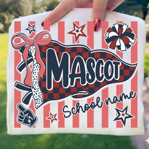May include: A white t-shirt featuring a graphic print with vertical coral stripes. The design includes a dark blue pennant with a red and black checkered pattern, the text MASCOT and School name, a coral bow, a cheerleader pom-pom, stars, and a foam finger.