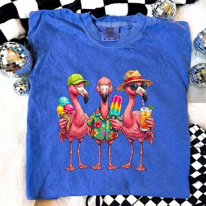 May include: A blue Comfort Colors t-shirt featuring a graphic of three pink flamingos. One flamingo wears a green cap and holds an ice cream cone, another holds a popsicle, and the third wears a straw hat and sunglasses, sipping a drink.