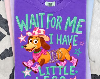 Può includere: Una t-shirt viola a maniche corte con la grafica di un bassotto marrone dal corpo a molla, che indossa un cappello da cowboy rosa, una bandana rosa e stivali rosa. Il testo recita WAIT FOR ME I HAVE LITTLE LEGS in lettere verde chiaro.