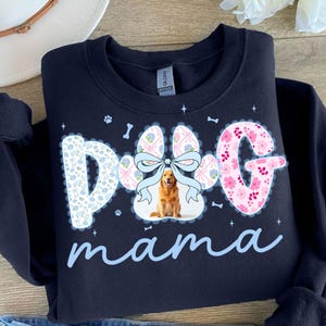 May include: Navy blue sweatshirt with a paw print design that says "Dog mama". The paw print is decorated with a bow and a golden retriever image. The letters are decorated with floral patterns.