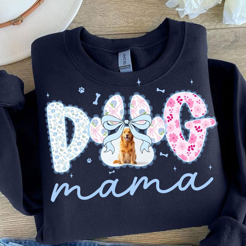 May include: Navy blue sweatshirt with a paw print design that says "Dog mama". The paw print is decorated with a bow and a golden retriever image. The letters are decorated with floral patterns.
