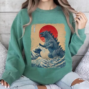 May include: A seafoam green sweatshirt with a Godzilla graphic in a Japanese art style. The design features a red sun, ocean waves, and the word "Kaiju" in Japanese characters. The graphic is printed on the front of the sweatshirt.
