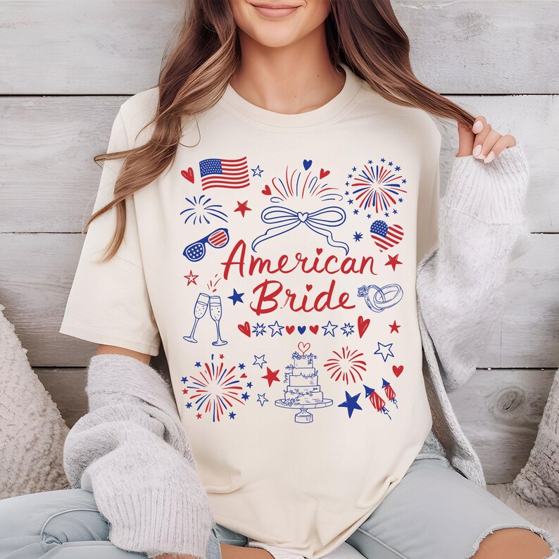 May include: Cream-colored t-shirt with a patriotic design featuring the text "American Bride" in red script. The design includes fireworks, hearts, stars, an American flag, sunglasses, champagne glasses, and a wedding cake.