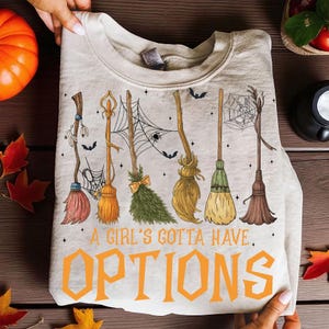 May include: A beige sweatshirt featuring a Halloween-themed design. The design includes various broom illustrations, spiderwebs, and bats. The text "A Girl's Gotta Have Options" is printed in orange.