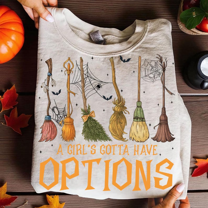 May include: A beige sweatshirt featuring a Halloween-themed design. The design includes various broom illustrations, spiderwebs, and bats. The text "A Girl's Gotta Have Options" is printed in orange.
