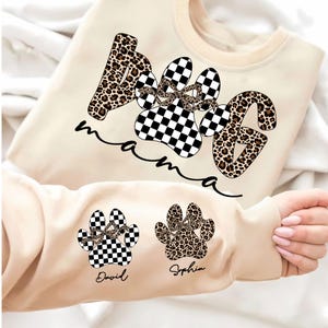 May include: Cream-colored sweatshirt with the words "Dog Mama" in a paw print design. The paw prints are a mix of leopard print and black and white checkered patterns. The sleeve features two additional paw prints with names.