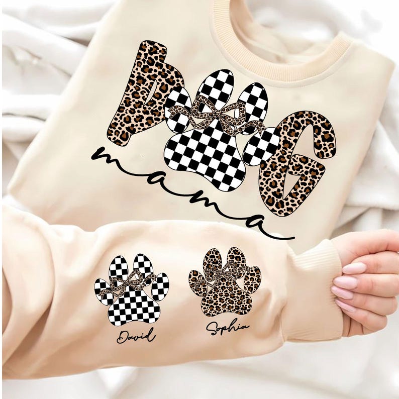 May include: Cream-colored sweatshirt with the words "Dog Mama" in a paw print design. The paw prints are a mix of leopard print and black and white checkered patterns. The sleeve features two additional paw prints with names.