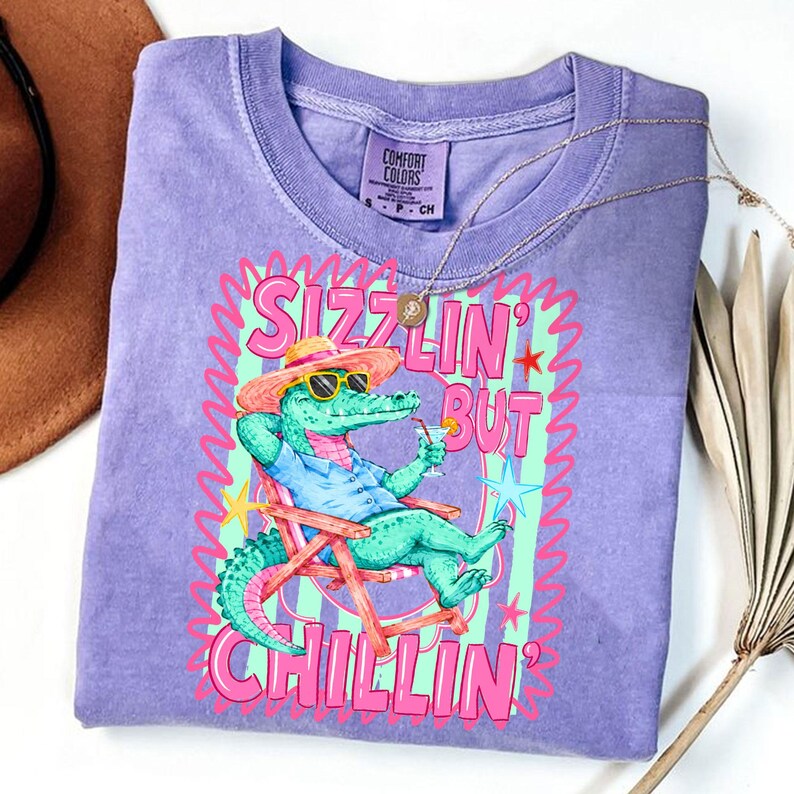 May include: A purple Comfort Colors t-shirt featuring a cartoon alligator in sunglasses and a straw hat, relaxing in a beach chair with a drink. The shirt has the text "Sizzlin' But Chillin'" in pink and green.