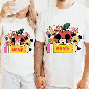 May include: White t-shirts with a colorful school-themed graphic. The design features a red apple with a Mickey Mouse face, pencils, a school bus, and the word "NAME" on a yellow pencil.
