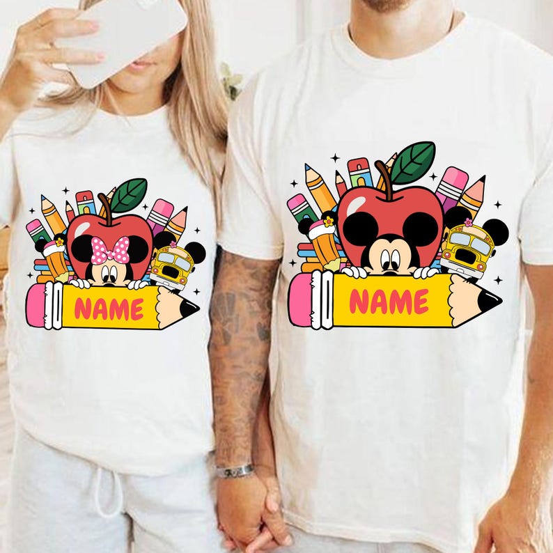 May include: White t-shirts with a colorful school-themed graphic. The design features a red apple with a Mickey Mouse face, pencils, a school bus, and the word "NAME" on a yellow pencil.