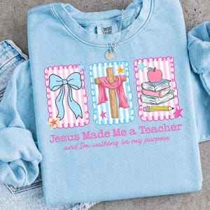 May include: A light blue sweatshirt featuring a graphic with three scalloped rectangles containing a blue bow, a wooden cross draped with a pink cloth, and a stack of books with an apple and pencil. Text reads: Jesus Made Me a Teacher and I'm walking in my purpose.