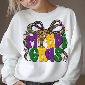 May include: White sweatshirt with a colorful Mardi Gras design. The graphic features the words "Mardi Gras" in purple, yellow, and green, with a bow and a crawfish in the center. The design includes stars and fleur-de-lis.