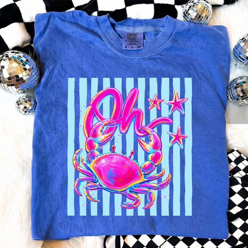 May include: A blue Comfort Colors t-shirt featuring a vibrant graphic of a crab with the word "Oh" in pink. The crab is set against a light blue striped background with pink stars. The shirt is a size small.