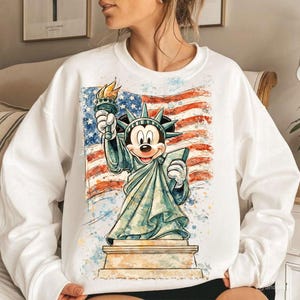 May include: White sweatshirt featuring a graphic of Mickey Mouse as the Statue of Liberty, holding a torch and tablet, set against a watercolor American flag. The design is a patriotic and whimsical blend of iconic symbols.