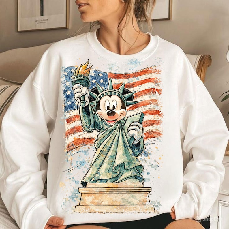 May include: White sweatshirt featuring a graphic of Mickey Mouse as the Statue of Liberty, holding a torch and tablet, set against a watercolor American flag. The design is a patriotic and whimsical blend of iconic symbols.