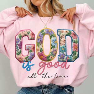 Op de afbeelding: Een lichtroze sweatshirt met de tekst "GOD is good all the time". De letters "GOD" zijn versierd met kleurrijke bloemen. Het woord "is" is geschreven in lichtblauw.