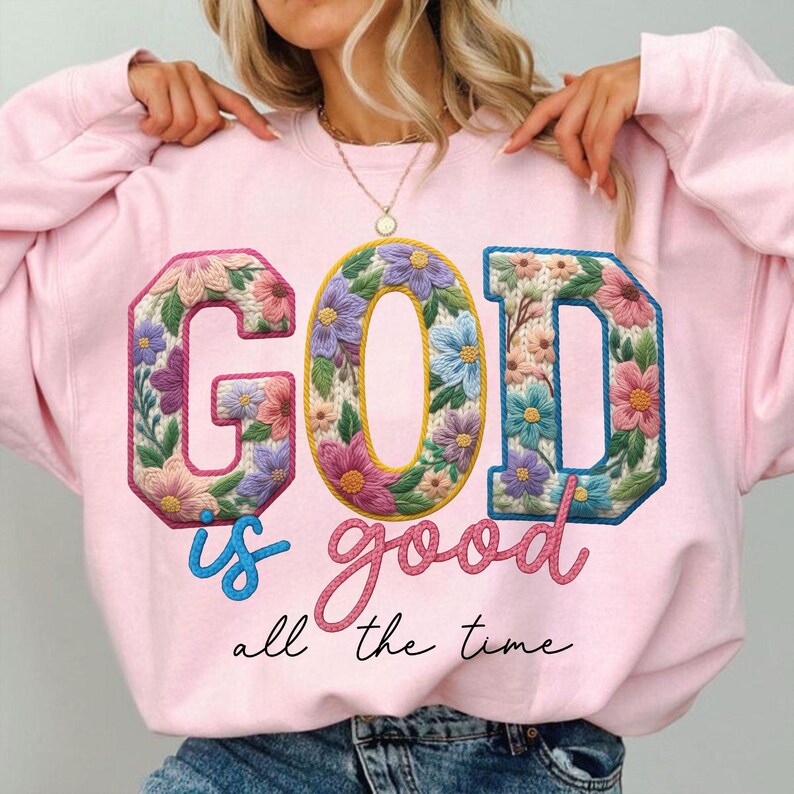 Op de afbeelding: Een lichtroze sweatshirt met de tekst "GOD is good all the time". De letters "GOD" zijn versierd met kleurrijke bloemen. Het woord "is" is geschreven in lichtblauw.