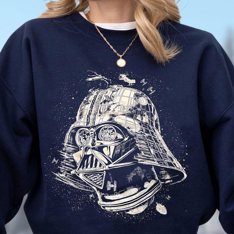 May include: Navy blue sweatshirt featuring a detailed white graphic of Darth Vader's helmet. The design includes elements of space and starships. The sweatshirt is a crew neck style, perfect for casual wear.