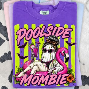 May include: Purple t-shirt with a graphic of a ghost-like figure wearing sunglasses and a leopard print headband, lounging on a pink flamingo float. The text "Poolside Mommie" is displayed in pink. The design includes bats and a cocktail.