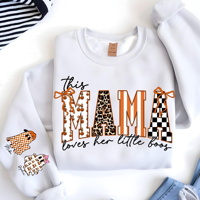 May include: White sweatshirt with the text "This MAMA loves her little boos" in black script. The word "MAMA" is in orange, leopard print, and black and white checkered patterns. Two ghost graphics are on the sleeve.