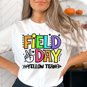 May include: White t-shirt with a colorful graphic that says "Field Day" with a peace sign and an arrow pointing to the right that says "Yellow Team".
