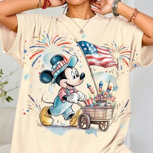 May include: Cream-colored t-shirt featuring a cartoon Mickey Mouse character dressed in patriotic attire, pulling a cart filled with fireworks and American flags. The design includes a waving American flag and festive fireworks in red, white, and blue.