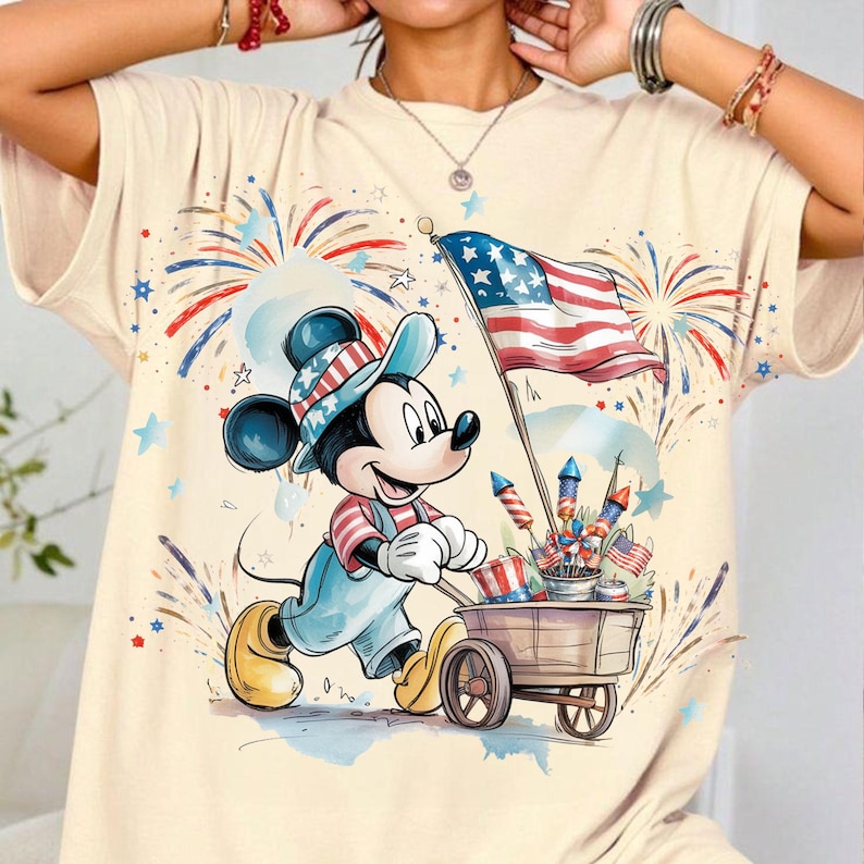 May include: Cream-colored t-shirt featuring a cartoon Mickey Mouse character dressed in patriotic attire, pulling a cart filled with fireworks and American flags. The design includes a waving American flag and festive fireworks in red, white, and blue.