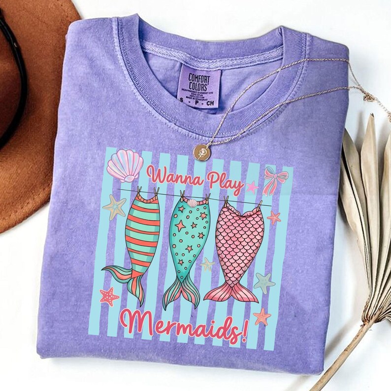 May include: A lilac Comfort Colors t-shirt featuring a graphic of three mermaid tails hanging on a clothesline. The text "Wanna Play Mermaids!" is printed below the tails. A gold necklace and a brown hat are visible.