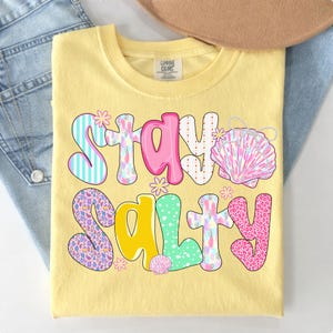 May include: Yellow t-shirt with the words "Stay Salty" in a colorful, whimsical font. The design includes seashells and floral accents. The shirt is folded on top of a pair of blue jeans.