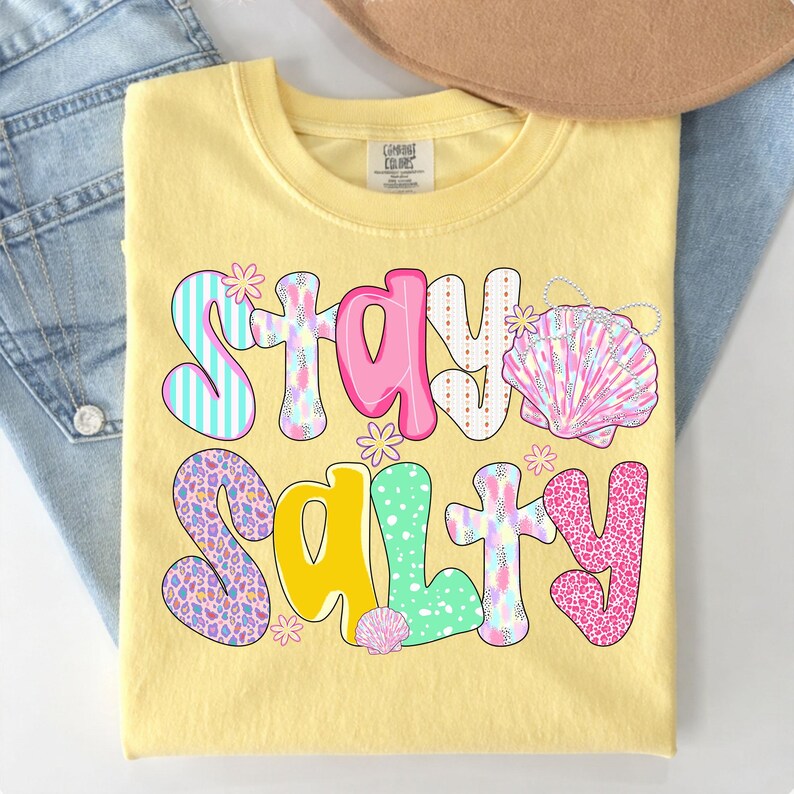 May include: Yellow t-shirt with the words "Stay Salty" in a colorful, whimsical font. The design includes seashells and floral accents. The shirt is folded on top of a pair of blue jeans.