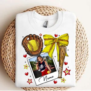 May include: White t-shirt with a softball theme. It features a baseball bat, a glove with a softball, a yellow bow, and a photo of a girl playing softball. The text "Name" is printed on the photo.