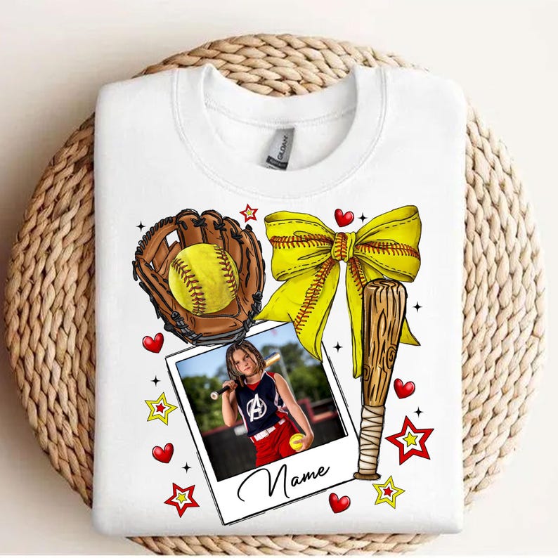 May include: White t-shirt with a softball theme. It features a baseball bat, a glove with a softball, a yellow bow, and a photo of a girl playing softball. The text "Name" is printed on the photo.