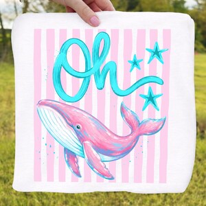 May include: White square towel with a pink and blue whale illustration. The whale is set against a background of pink vertical stripes. The word "Oh" is written in turquoise script above the whale, with three turquoise stars.