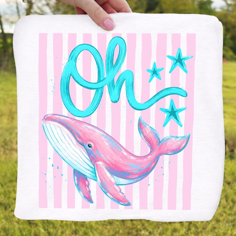 May include: White square towel with a pink and blue whale illustration. The whale is set against a background of pink vertical stripes. The word "Oh" is written in turquoise script above the whale, with three turquoise stars.
