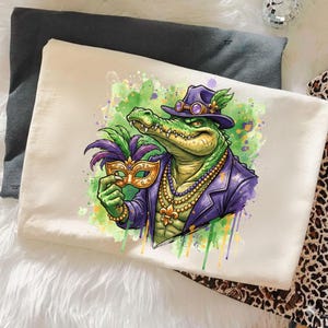 May include: Cream-colored t-shirt featuring a cartoon alligator in a purple suit and hat, holding a gold mask with purple and green feathers. The alligator wears a pearl necklace and has green and yellow accents.