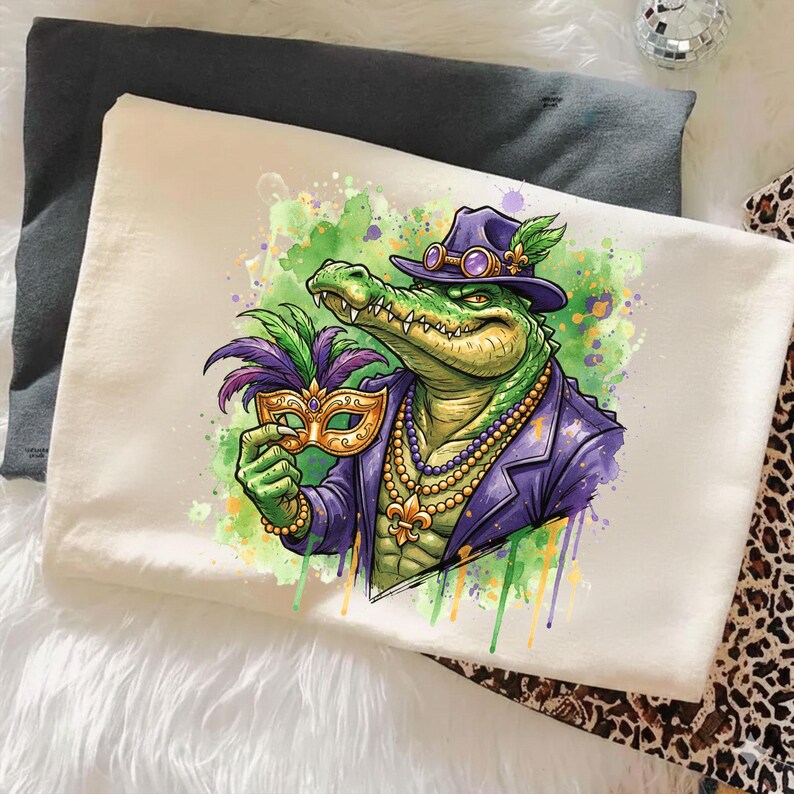 May include: Cream-colored t-shirt featuring a cartoon alligator in a purple suit and hat, holding a gold mask with purple and green feathers. The alligator wears a pearl necklace and has green and yellow accents.