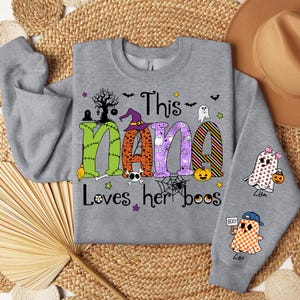 May include: A gray sweatshirt with the text "This Nana Loves her boos" in colorful Halloween-themed letters. The design includes ghosts, pumpkins, and a witch hat. The sleeves feature ghost graphics.