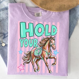 May include: A light purple t-shirt with the words "HOLD YOUR" in turquoise block letters. Below the text is a brown horse with a saddle and bridle. Pink vertical stripes and blue stars are behind the horse.