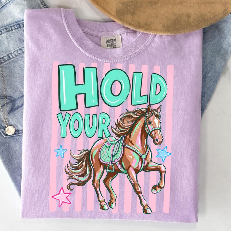 May include: A light purple t-shirt with the words "HOLD YOUR" in turquoise block letters. Below the text is a brown horse with a saddle and bridle. Pink vertical stripes and blue stars are behind the horse.