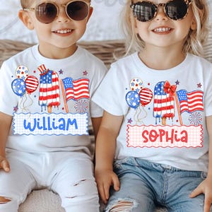 May include: Two white t-shirts with patriotic designs. One shirt has the name "William" and the other has "Sophia". Each shirt features a popsicle, balloons, and an American flag. The popsicle design has red, white, and blue colours.