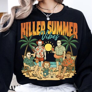 May include: Black sweatshirt with a graphic featuring horror movie characters on a beach, palm trees, and the text "KILLER SUMMER Vibes" in orange and blue. The design includes a sun and sandcastles.