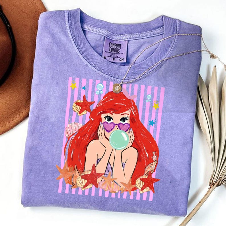 May include: A lilac Comfort Colors t-shirt with a cartoon mermaid graphic. The mermaid has red hair, heart-shaped sunglasses, and a bubble. The design includes pink stripes, starfish, and seashells. A gold necklace is draped over the shirt.