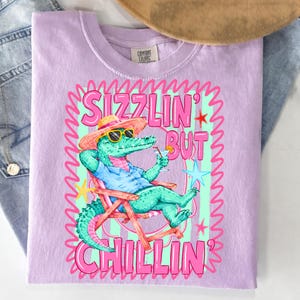May include: A light purple t-shirt with a graphic of an alligator wearing sunglasses and a hat, relaxing in a beach chair with a cocktail. The text reads "Sizzlin' But Chillin'" in pink and green. The shirt is made of a soft material.