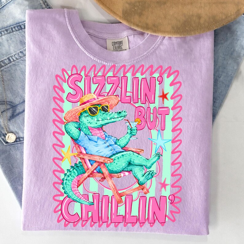 May include: A light purple t-shirt with a graphic of an alligator wearing sunglasses and a hat, relaxing in a beach chair with a cocktail. The text reads "Sizzlin' But Chillin'" in pink and green. The shirt is made of a soft material.