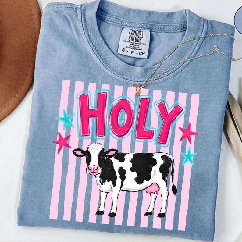 Puede incluir: Camiseta Comfort Colors azul claro con la palabra "HOLY" en rosa con contorno azul, estrellas rosas y azules, y un gr&aacute;fico de vaca en blanco y negro. La camiseta tiene cuello redondo y mangas cortas.